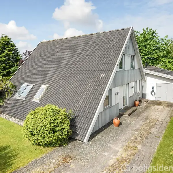 A-formet hus med sort tegltag står på en velplejet grøn græsplæne. Flere buske omgiver indkørslen med grus og en tilhørende garage. Skyfri himmel i baggrunden.