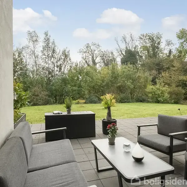 Sofa og stole står på en flisebelagt terrasse, hvor de omkranser et bord med pynt. Omkring terrassen er der en græsplæne og frodig bevoksning under en blå himmel.