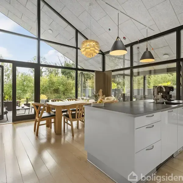 Et moderne køkken-alrum med store vinduer. Et køkkenbord i forgrunden, spisebord og stole bagved. Højt til loftet med hængelamper. Udenfor ses en terrasse og grøn natur.