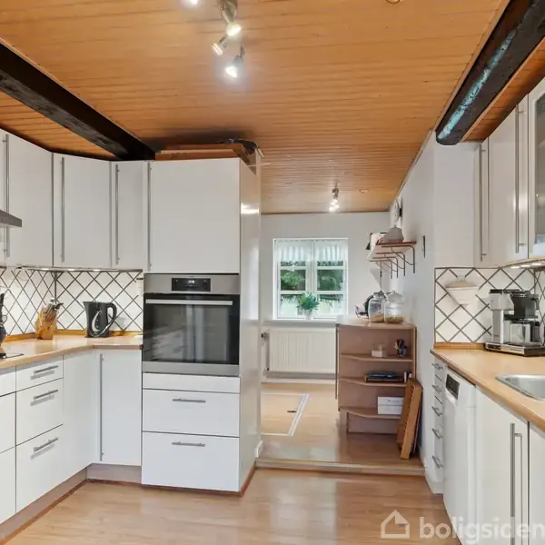 Et moderne køkken med hvide skabe er organiseret med indbyggede apparater og en vask. Træloftet og flisebeklædningen giver en varm atmosfære i rummet, der har udsigt til haven.