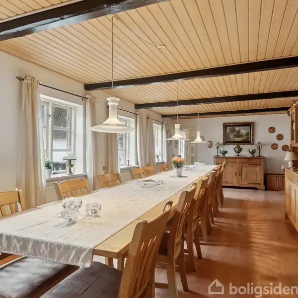 Langt spisebord med hvid dug og træsæder omgivet af stole i et lyst rum med træloft. Væghængte lamper over bordet, skænk og skabe langs væggen.