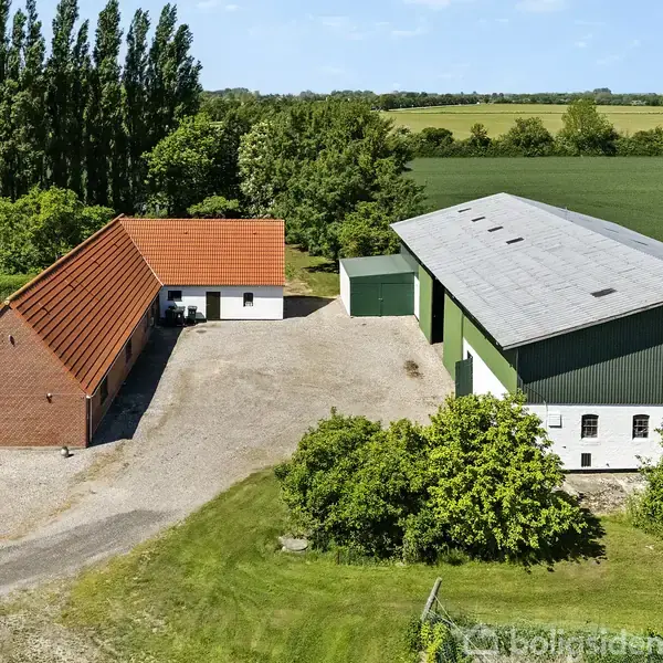 En gård med en rødtagt bygning og en stor lade står stille i et landligt område omgivet af marker og træer under en blå himmel.