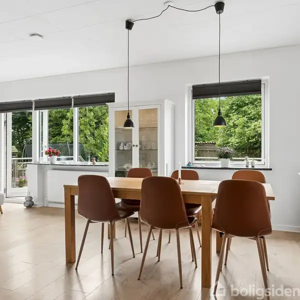 Spisebord med seks stole, omgivet af store vinduer, slipper naturligt lys ind i det åbne, moderne køkken-alrum med minimalistisk indretning og udsigt til en grøn have.