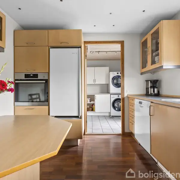 Blomster på et køkkenbord ved siden af beige træskabe og køkkenmaskiner. Dør fører til vaskerum med hvide skabe og vaskemaskine.