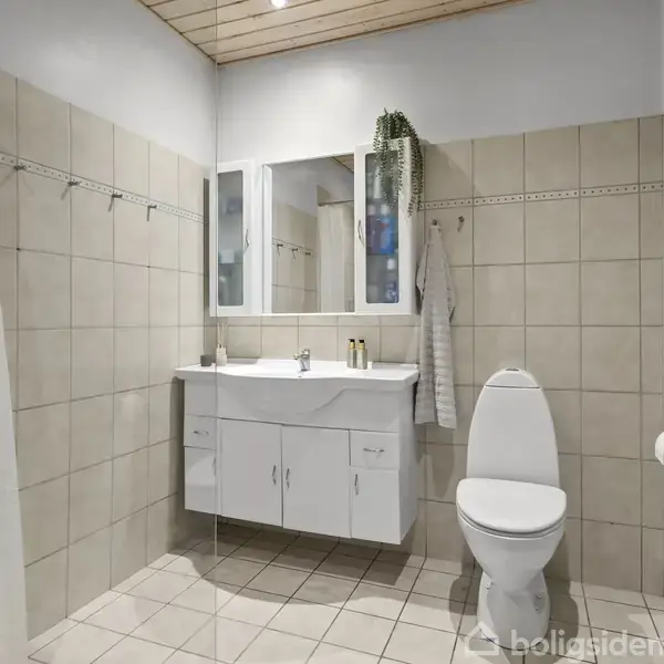 Badeværelse med hvidt toilet og vaskeskab, omkranset af lyse fliser. En håndvask er placeret under et spejl. Der hænger et håndklæde ved siden af.