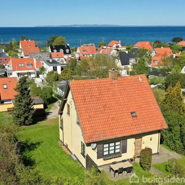 Et gult hus med rødt tegltag ligger i en grøn have. I baggrunden ses en kystby med flere huse og en udsigt til havet under en blå himmel.