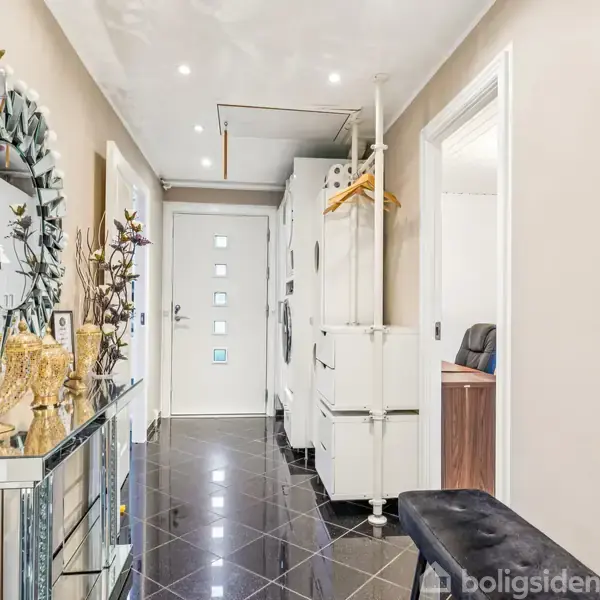 Der er en lys gang med sorte flisegulve, en dekorativ spejl på væggen og diverse pyntegenstande. Der er et smalt bord til højre, og døren for enden har små vinduer.