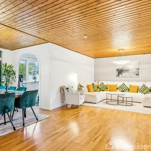 En moderne stue med en stor hvid sofa fyldt med farverige puder. Til venstre står et spisebord med grønne stole. Rummet har et træloft og lyse trægulve.