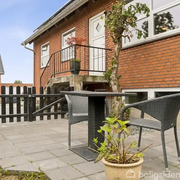To sorte stole står ved et kvadratisk bord på en flisebelagt terrasse foran et rødt murstenshus. Ved siden af er der et lille hegn og en plantet krukke.