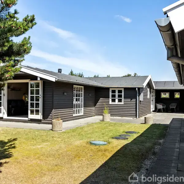 Et mørkt træsommerhus med åbne franske døre leder til en grøn have. Til venstre ses et træ, til højre en overdækket gang. Klar himmel i baggrunden.