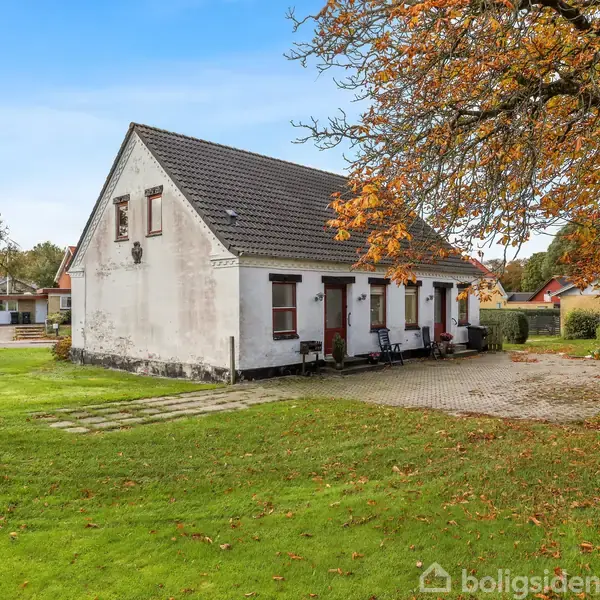 Et hvidt hus med sort tag står roligt på en grøn græsplæne. Foran huset er et brolagt område og et stort træ med efterårsblade.