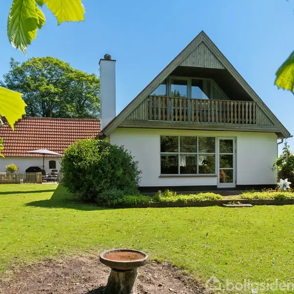 Et hvidt hus med trekantet tag og altan står i en frodig have. Græsplænen er omkranset af buske og træer, og foran er en fuglebad. Tekst: "Boligsiden".