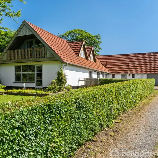 Et hvidt hus med rødt sadeltag står i en frodig have, omgivet af grønne buske. En grusvej løber ved siden af huset under en klar blå himmel.