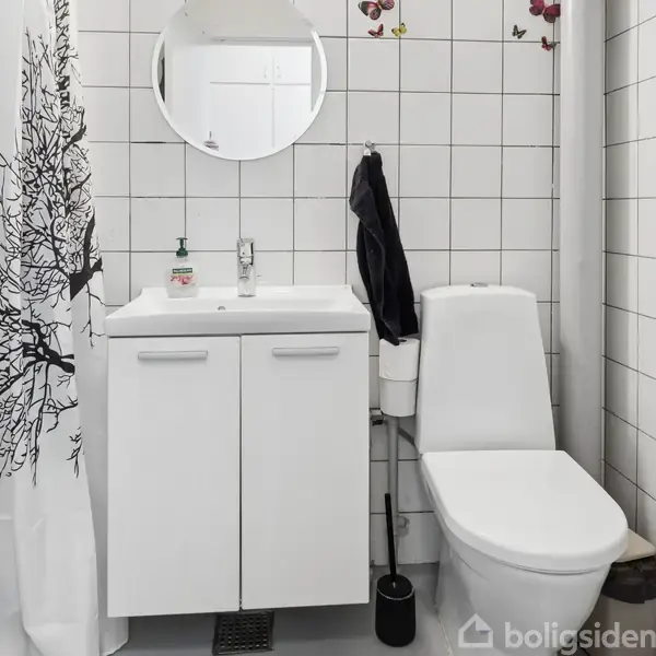 Et badeværelse med et toilet ved siden af et hvidt skab med håndvask, spejl over vasken, og sort/hvidt bruseforhæng. Væggene er dækket af hvide fliser.