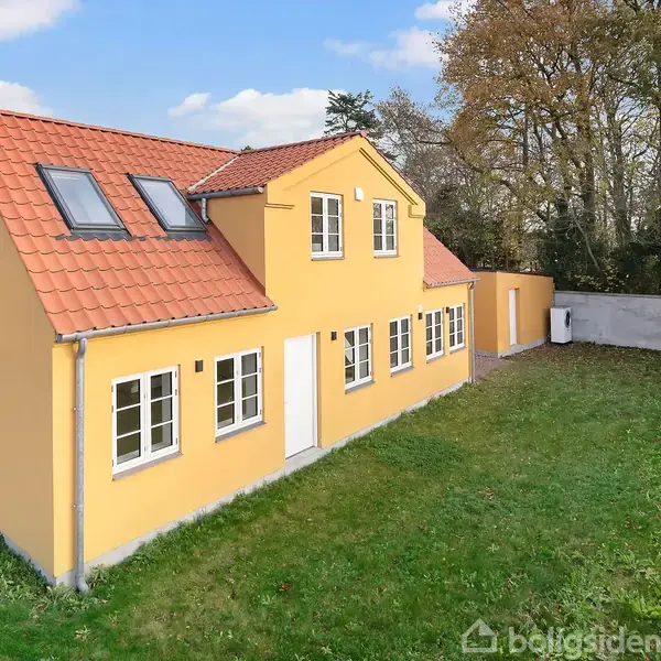 Et gult hus med rødt tag står på en græsplæne. Der er flere vinduer, nogle ovenlysvinduer, og det ligger tæt på et skovområde med træer i baggrunden.