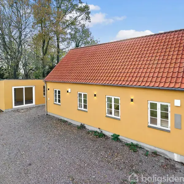 Et gult hus med rødt tegltag står stille på en grusbelagt indkørsel omgivet af træer, med flere vinduer og en tilstødende moderne, rektangulær tilbygning.