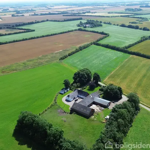 Luftfoto af en gård med flere bygninger omgivet af grønne marker og træer i et landligt område. Veje snor sig gennem landskabet, der er opdelt i forskellige jordlodder.