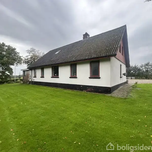 Et hvidt hus med skråt tag står på en grøn græsplæne omgivet af træer under en overskyet himmel.