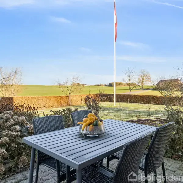 Et havebord i metallisk finish står i en terrasse med fire stole omkring det. På bordet er en skål med frugt. I baggrunden ses en flagstang og en hæk, omgivet af grønne marker.
