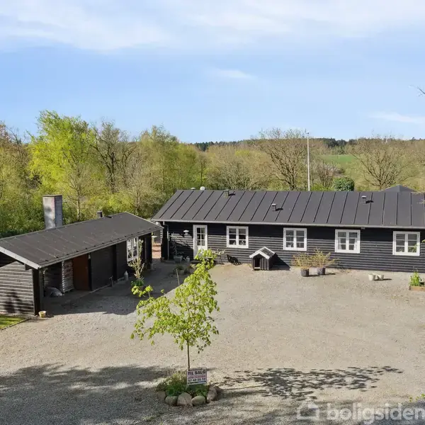 En mørk træbygning med sort tag sidder stille i naturen omgivet af træer, med en grusvej foran og en garagebygning til venstre.