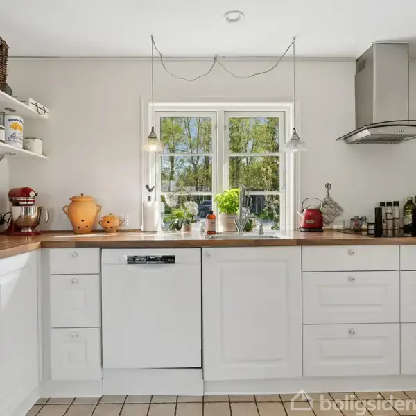 Hvidt køkken med træbordplade viser opvaskemaskine under vindue. Køkkenredskaber og krydderier står på hylder. Keramik og brødkurv pynter. Grønne træer sees udenfor vinduet.