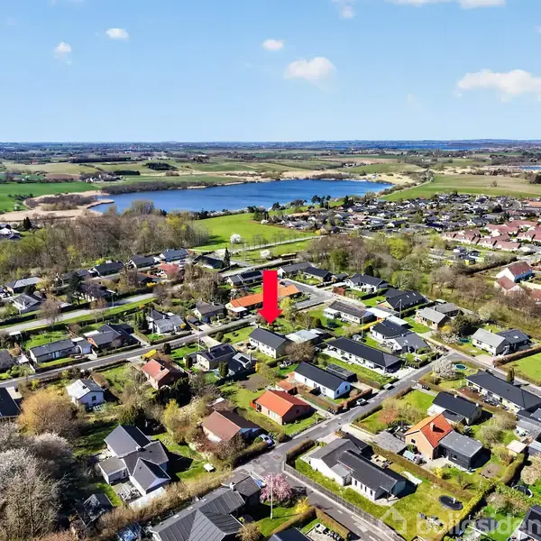 Et moderne køkken med hvide og sorte skabe; en køkkenø fungerer som siddeområde med to høje stole og dekoreret med frugtskål og plante. Store vinduer giver naturligt lys.