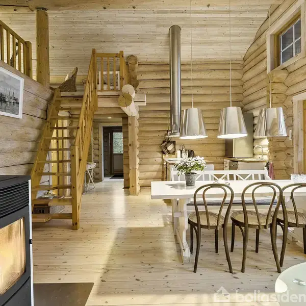 Brændeovn står i en stue med træbjælkevægge, spiraltrappe til loftet, lang spisebord med stole, og moderne lamper hænger fra loftet. Store vinduer viser skov udenfor.