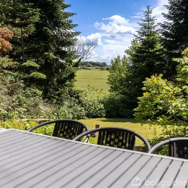 Et udendørs bord med sorte stole står på en terrasse. Bagved er en grøn have med buske og høje træer, efterfulgt af et åbent landskab under en skyet, blå himmel.