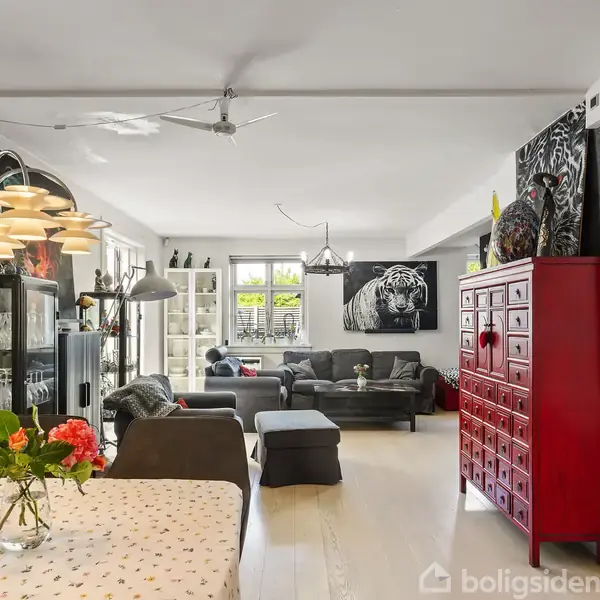En stue med spisebord og vase med blomster. Sofaer står ved væggene med tigerkunst. En stor, rød kommode er nær væggen. Rummet er lyst med store vinduer.