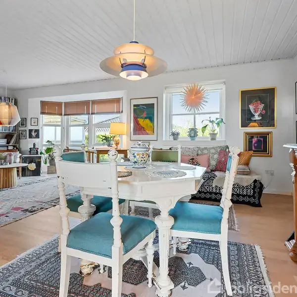 Et rundt spisebord med blå polstrede stole står i en stue med lyse vægge, dekoreret med farverige malerier og planter. Reol og sofaer er synlige i baggrunden.