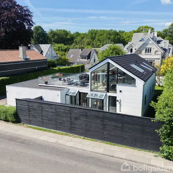 Et moderne hus med store glaspartier står bag et sort stakit. Huset har en terrasse på taget. Omgivet af andre huse og grønne træer på en villavej.