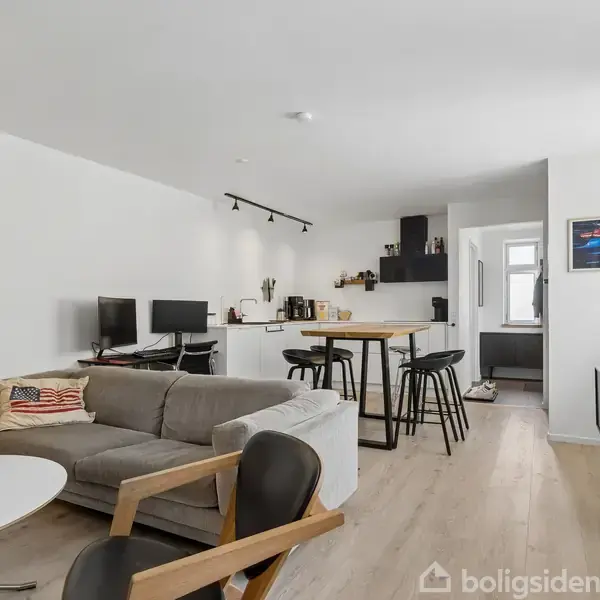 Stue med grå sofa og bord står mod en hvid væg. Der er to computere på et skrivebord. I baggrunden ses et åbent køkken med spisebord og tre stole.