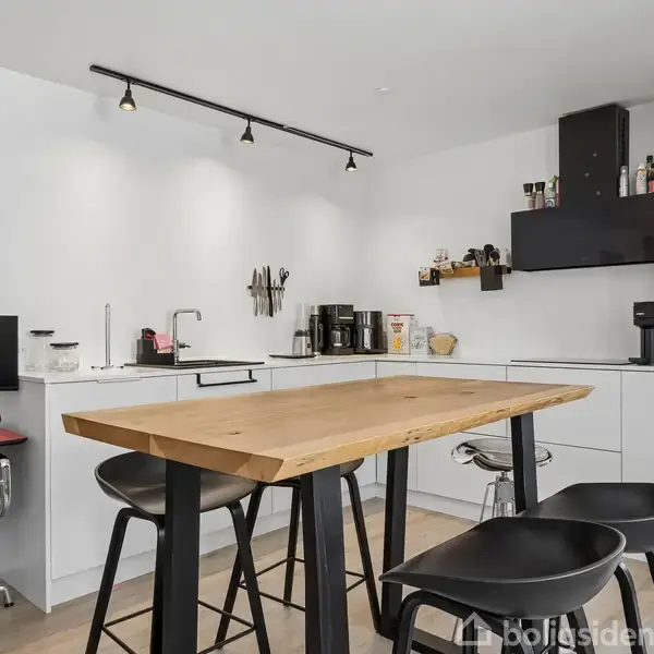 Et moderne køkken med et træbord omgivet af sorte stole dominerer rummet. Køkkenet har hvide skabe, sort emhætte, og der står køkkenapparater, som kaffemaskiner, på bordet. En computerplads ses til venstre.