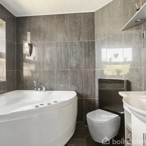 Badekar står stille i et moderne badeværelse med grå fliser, ved siden af et toilet og håndvask, mens et vindue med planter slipper lys ind.