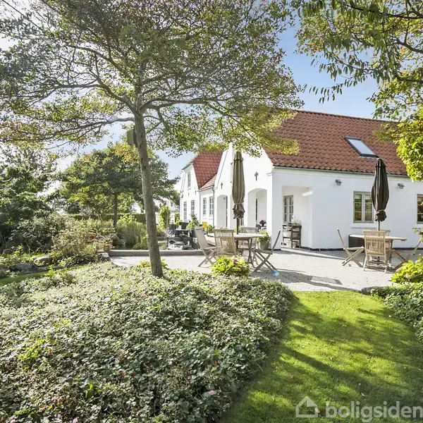 Hvidt hus med rødt tag står stille i en solrig have, omgivet af græs, træer og borde med parasoller i en frodig og rolig atmosfære.