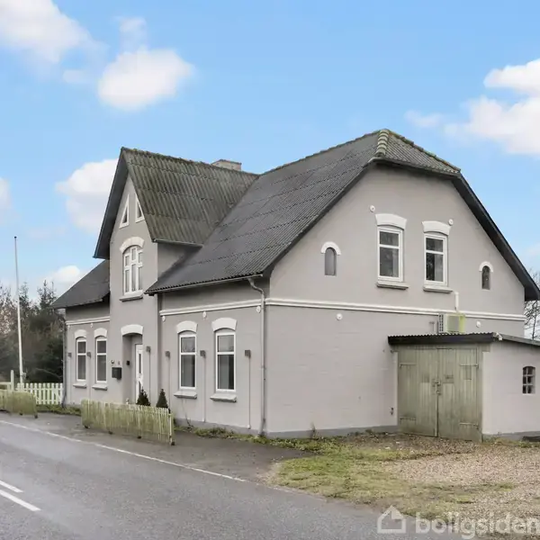 Huset står stille ved vejkanten, bygget i en klassisk stil med gråt facade og sort tag. Det er omgivet af en åben landevej og spredte træer under en blå himmel.