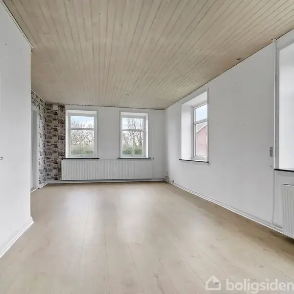 Et rummeligt, lyst rum med trægulve og træloft. To store vinduer på hver væg lader sollys strømme ind. Malede hvide vægge og en radiator pryder rummet.