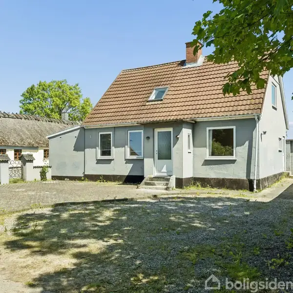Et gråt hus med rødt tag står stille i en solrig gårdsplads med grusbelægning. Til venstre ses en mindre bygning og træer omkranser området.