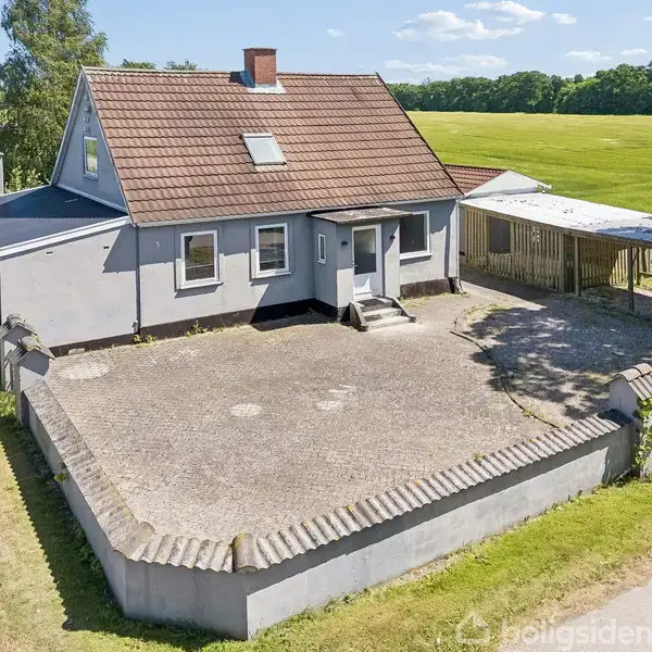 Et gråmalet hus med rødt tegltag står langs en landevej. Huset har en stor flisebelagt gårdsplads omkranset af en lav mur. Græsområder og træer omgiver ejendommen.