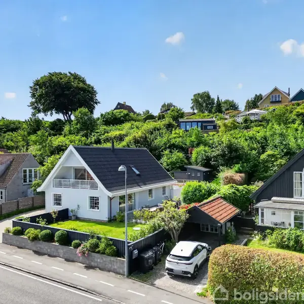 En hvid hus med sort tag har en velholdt have foran. To biler er parkeret i indkørslen. Omgivet af andre huse og træer i et grønt boligområde.