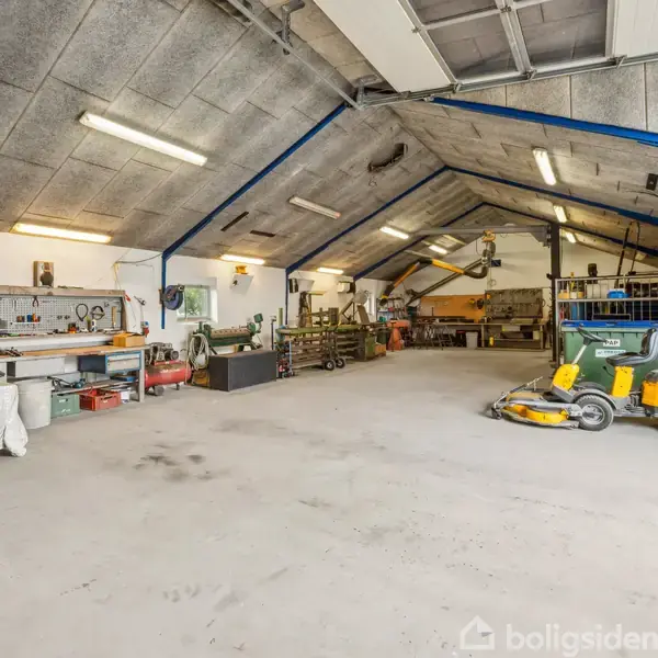 Et rummeligt værksted med en gul plæneklipper parkeret til højre. Værktøj og maskiner er organiseret langs væggene. Loftsbelysning oplyser det grå betongulv.