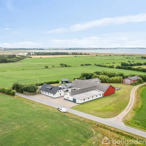 En gård beliggende i et åbent landskab udført i moderne stil med flere bygninger og en vej, der fører ind til ejendommen. Marker og skovområder omgiver stedet.