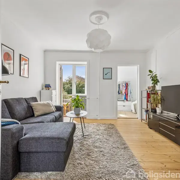 En stue med en grå sofa står ved siden af et sofabord. Fjernsynet står på en skænk. Rummet modtager lys fra en altandør, og et andet værelse ses i baggrunden.