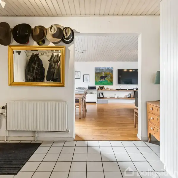 En entré med flisegulv viser jakker og hatte hængende ved et spejl. I baggrunden ses en stue med trægulv, møbler og kunst på væggene.