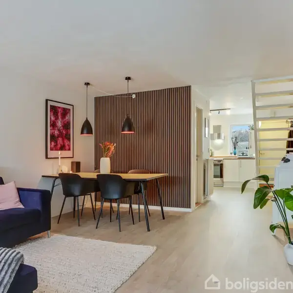 Stue med en mørk sofa, et tæppe og et rundt bord. Et spisebord med stole står nær skillevæg; trappe fører til øvre etage. Indretningen er moderne og lys.