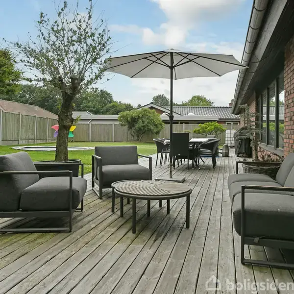 Træmøbler står på en terrasse med en parasol og et borde-sæt. I baggrunden er en have med et stakit, træer og græs. Solen skinner klart.