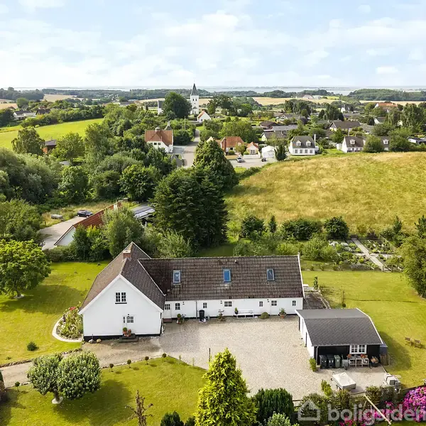 Hvidt hus med brunt tag ses fra luften. Det står i rolige, grønne omgivelser med en gårdsplads foran. Landskabet rundt er præget af marker og en lille landsby.