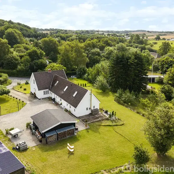 Hvidt hus med brun tag beliggende i frodigt, bakket landskab. Haven er stor med træer og græsplæne, omkranset af grønne områder og marker i baggrunden.