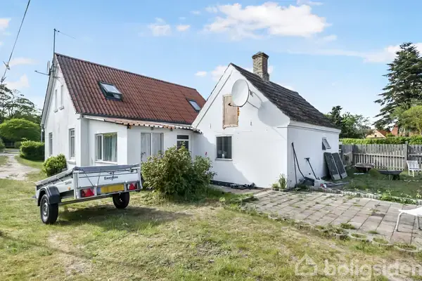 Huset har hvid facade og rødt tegltag, står på en græsplæne ved siden af en trailer. Der er en terrasse, og i baggrunden ses træer og en blå himmel.