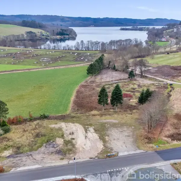 Landskab med åbent område ved siden af vejen, omgivet af marker og spredte træer. I baggrunden ses en sø med skovklædte bakker og en by på siden.